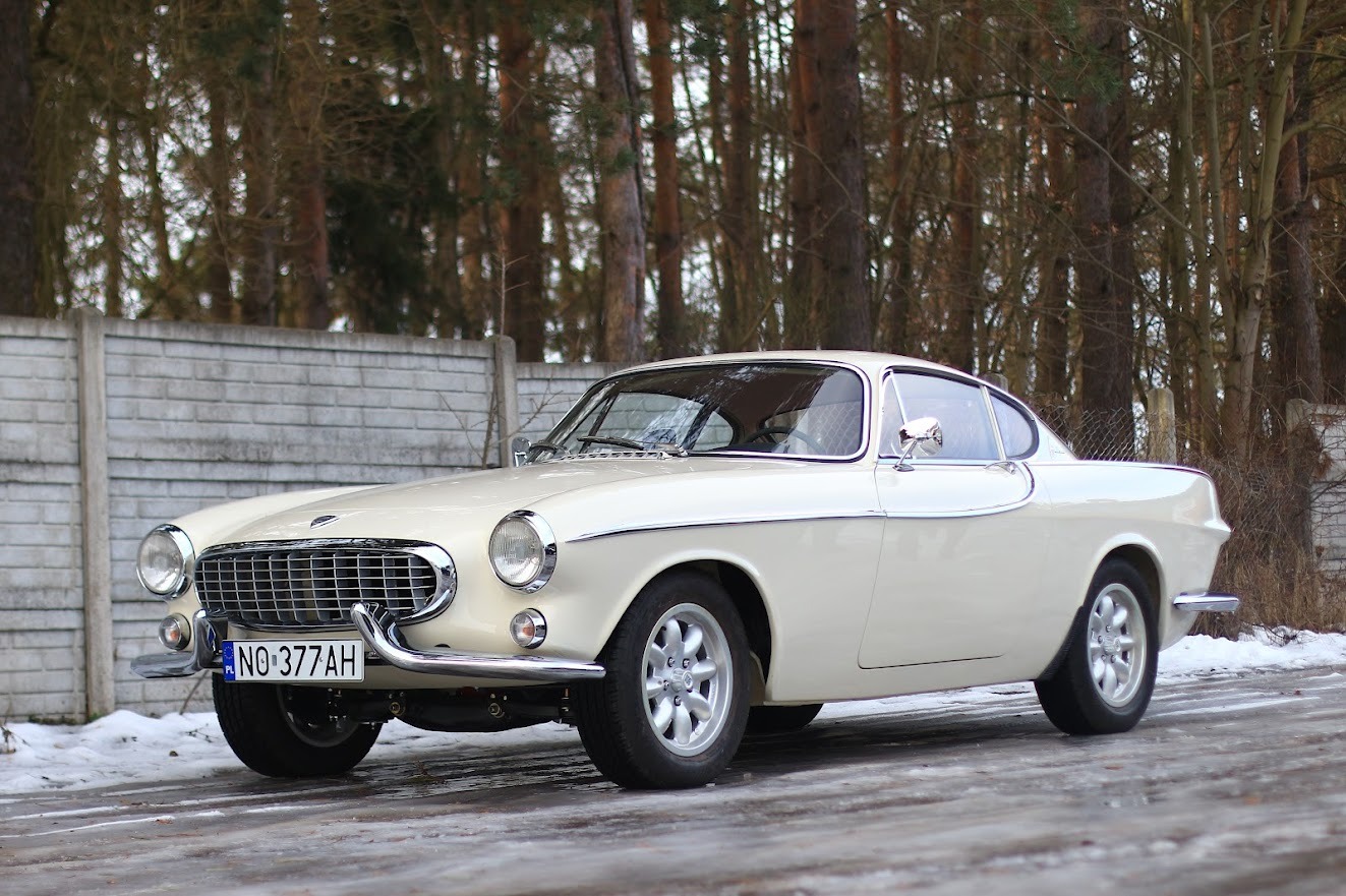 Białe zabytkowe Volvo P1800 z chromowanymi elementami i tablicą rejestracyjną na tle betonowego ogrodzenia i drzew