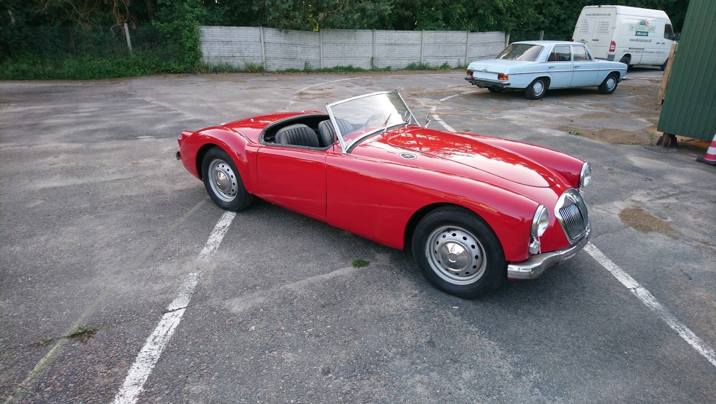 Czerwony, odrestaurowany kabriolet MGA z lat 50. na parkingu, obok zaparkowany jasnoniebieski Mercedes-Benz W114 oraz biały van z logotypem firmy zajmującej się renowacją pojazdów.