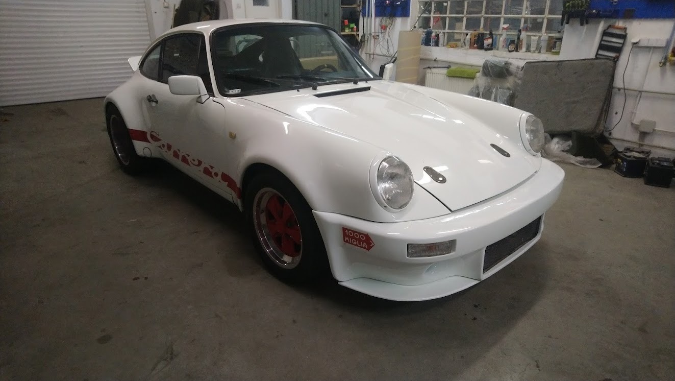 Białe Porsche 911 z czerwonymi akcentami i naklejką '1000 Miglia' na przednim zderzaku, stojące w warsztacie z widocznymi narzędziami i akcesoriami.