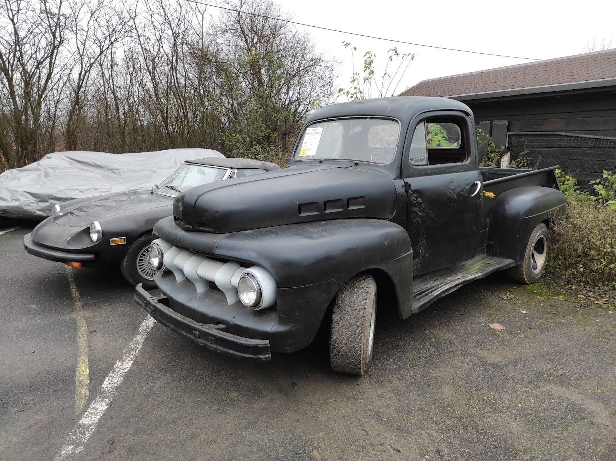 Czarna, matowa, klasyczna ciężarówka pickup z chromowanymi elementami grilla, zaparkowana obok czarnego sportowego samochodu i auta przykrytego plandeką.