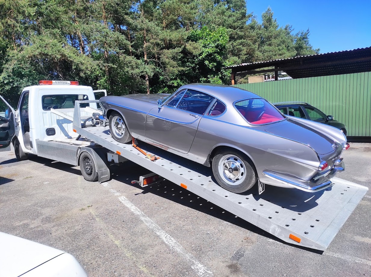Szary, klasyczny samochód Volvo P1800 z czerwonym wnętrzem transportowany na lawecie holowniczej. Widoczny fragment zielonego ogrodzenia w tle.