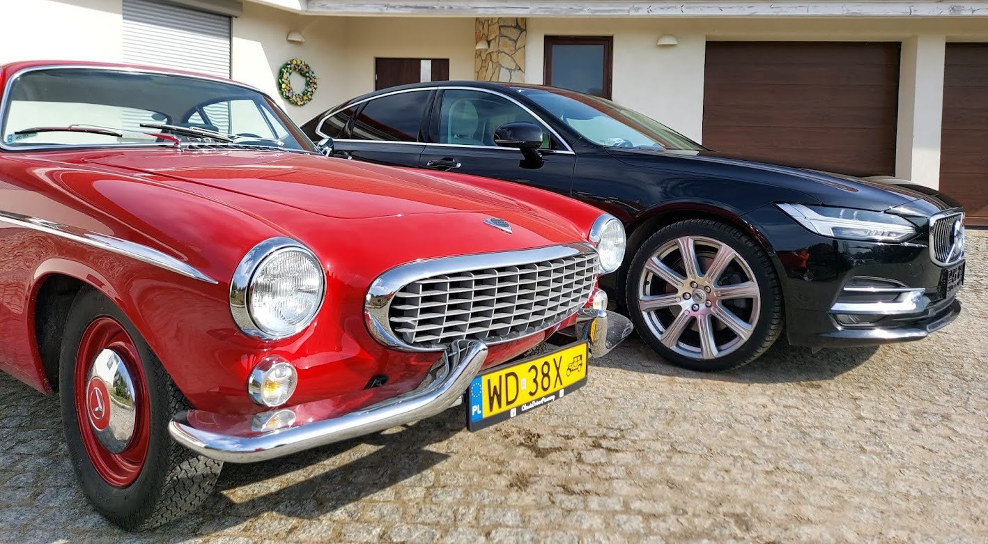 Czerwony, zabytkowy samochód Volvo P1800 z chromowanym grillem i żółtą tablicą rejestracyjną WD 38X, zaparkowany obok czarnego, nowoczesnego Volvo S90 na tle domu z garażem.