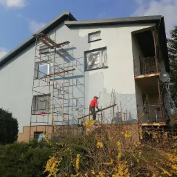 Dwukondygnacyjny dom w trakcie odświeżania elewacji, widoczne rusztowania i pracownik w czerwonej koszulce na wysokości