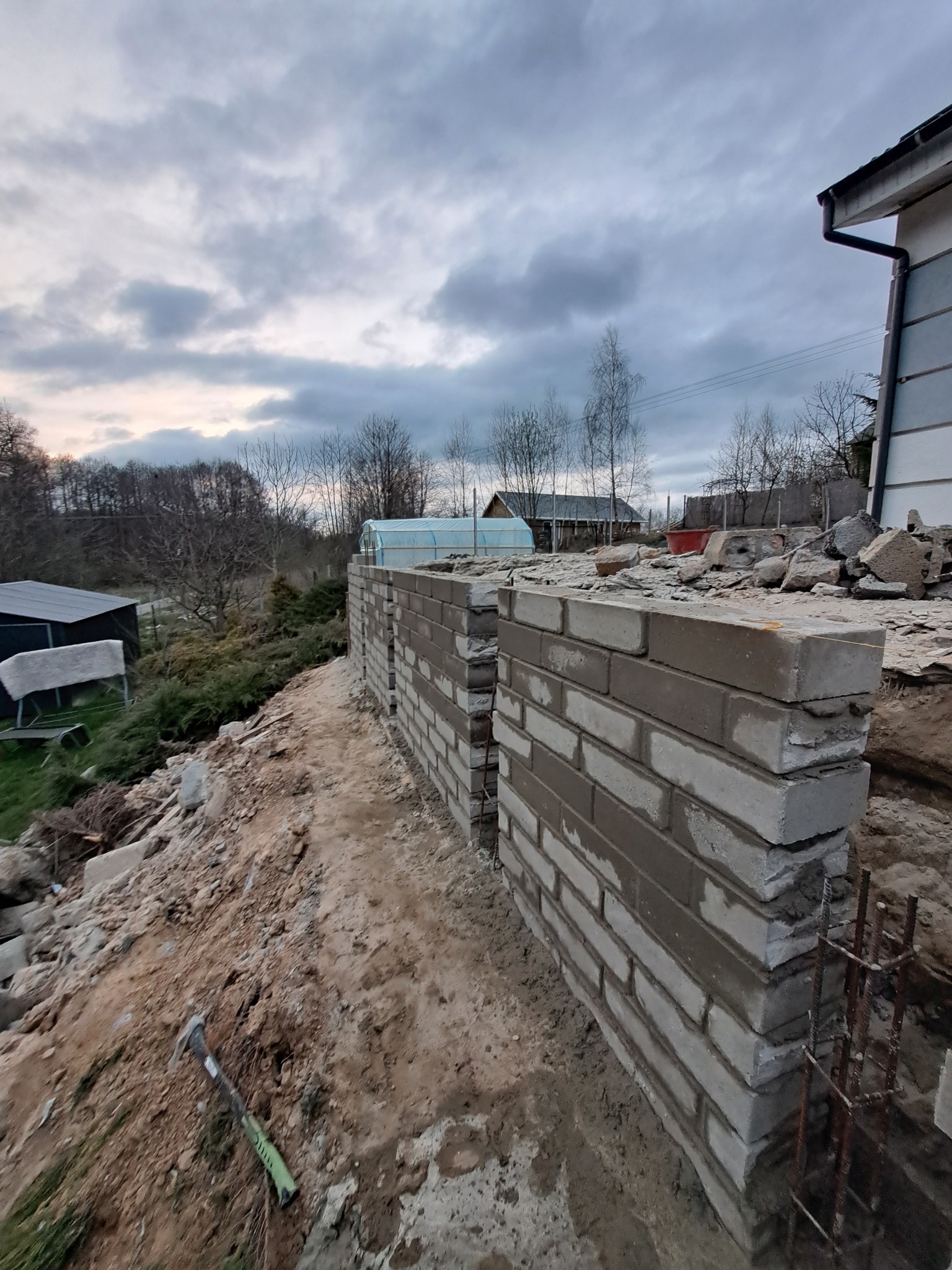 Wznoszenie muru z szarych bloczków betonowych na nierównym terenie, widoczne zbrojenie, w tle budynek mieszkalny i pochmurne niebo.