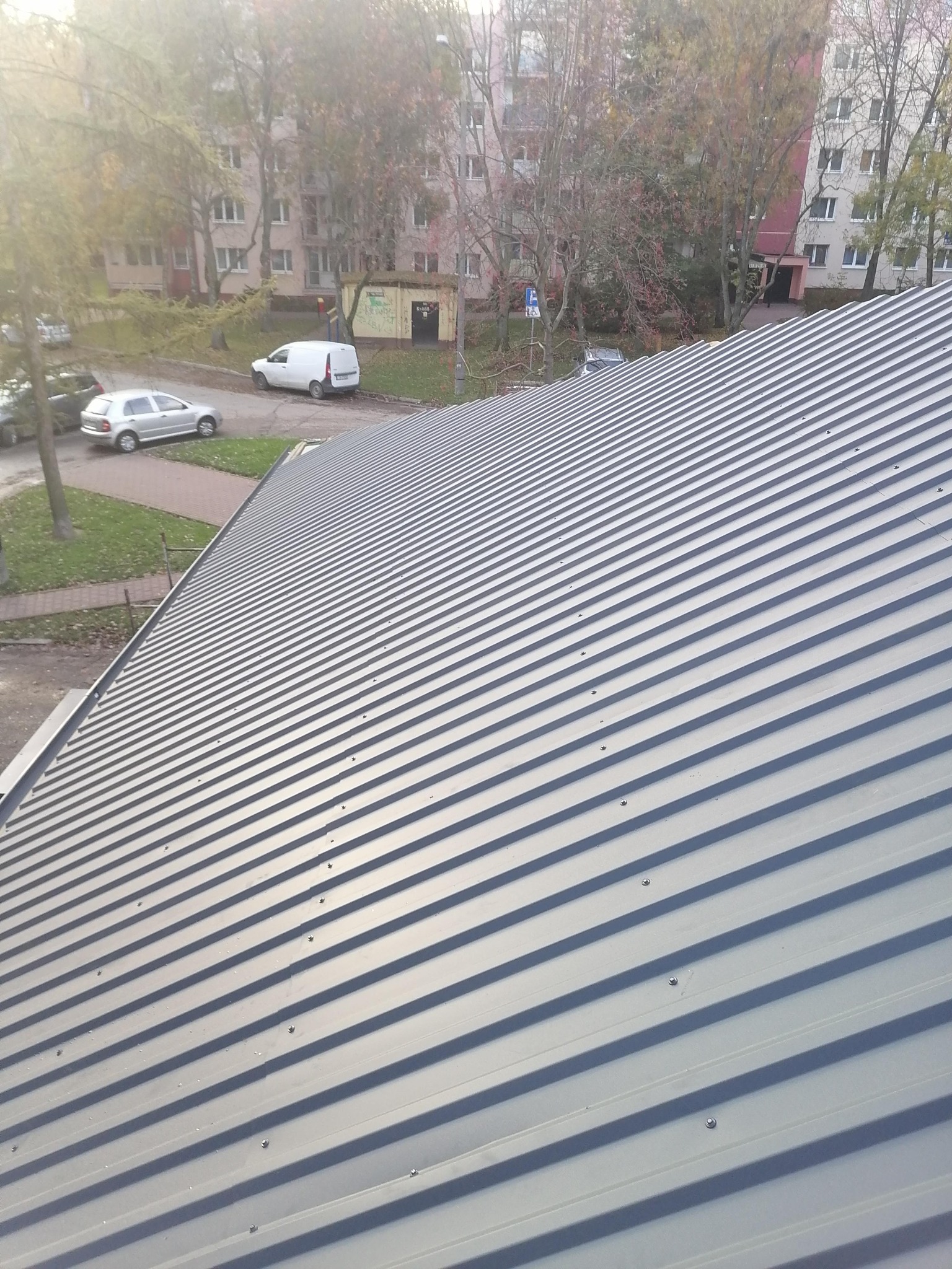 Nowe, szare blaszane pokrycie dachowe z widocznymi śrubami mocującymi, na tle budynków mieszkalnych i drzew w jesiennej scenerii.