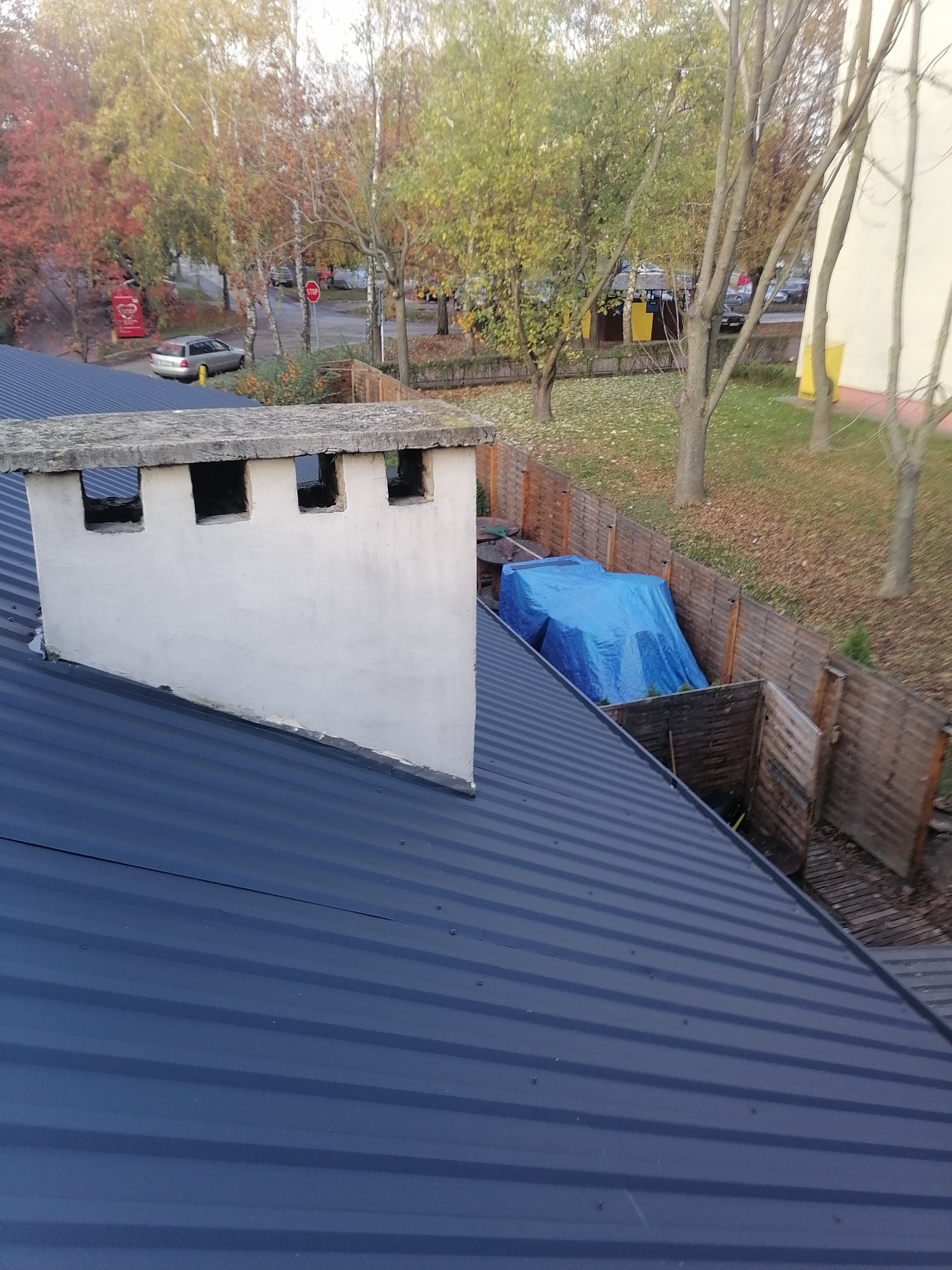 Niebieski dach z blachy trapezowej z białym kominem z czterema otworami, widok z góry na jesienne drzewa i zadaszony niebieską plandeką teren.
