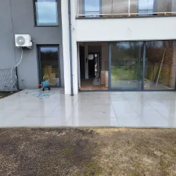 Świeżo ułożony, mokry taras z szarych płyt na zewnątrz nowoczesnego domu z widocznym wężem ogrodowym i przygotowanym podłożem pod trawnik.