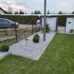 Ogród z trawnikiem, ścieżką z jasnego żwiru obramowaną kostką, niskimi krzewami i nowoczesnym ogrodzeniem z bramką, w tle żywopłot i dom.