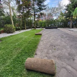 Rozwinięta i nierozwinięta trawa w rolkach na przygotowanym podłożu z siatką, widoczne drzewa i krzewy w ogrodzie.