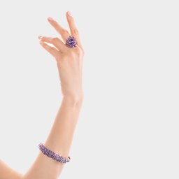 Adrianna Sołtys Fotografia - Delikatna dłoń modelki prezentuje fioletowy pierścionek i bransoletkę z kwiatowym motywem na tle jasnego studia. Elegancka biżuteria W.Kruk w zbliżeniu.