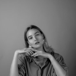Adrianna Sołtys Fotografia - Czarno-biały portret kobiety z blond włosami, w koszuli, z rękami pod brodą i dużymi kolczykami, siedzącej przy białym stole na neutralnym tle.