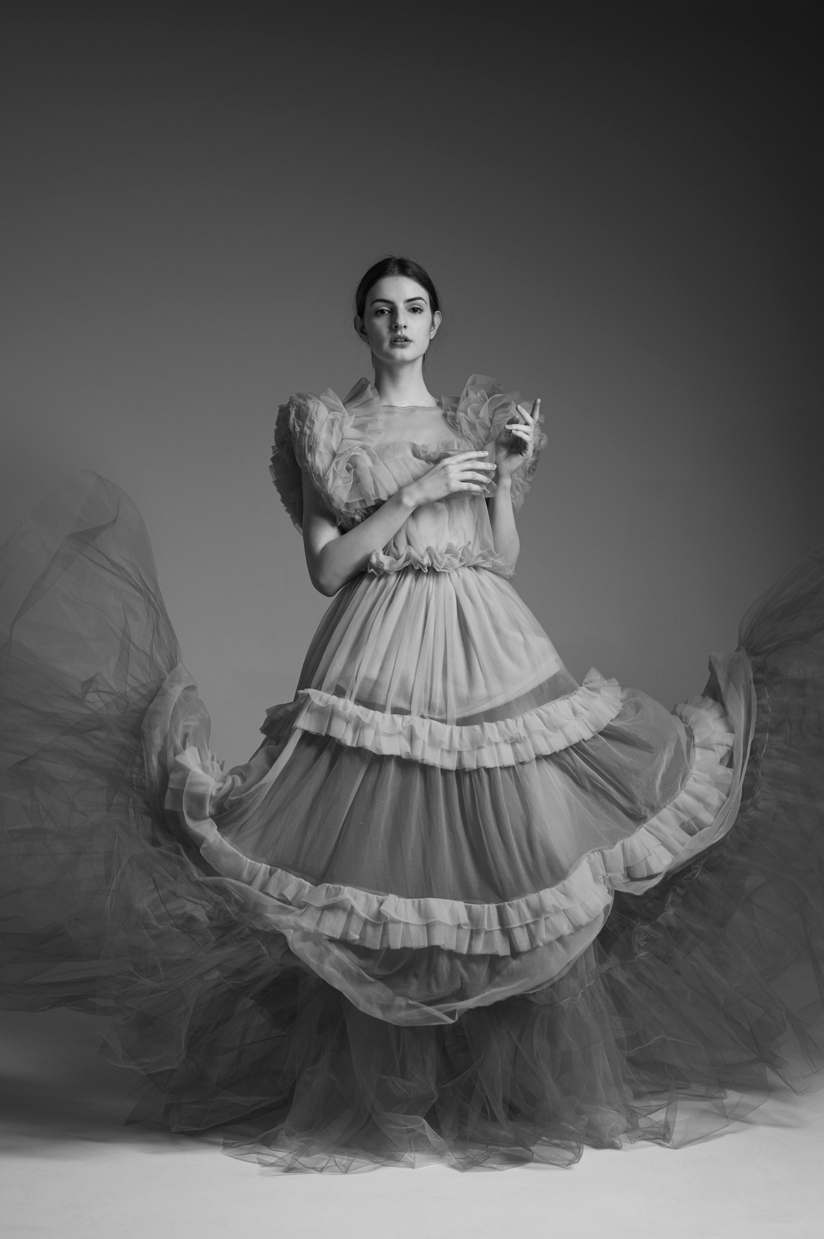 Portret kobiety w tiulowej sukni z falbanami, na szarym tle. Ujęcie studyjne, czarno-białe, z delikatnym światłem podkreślającym fakturę materiału.