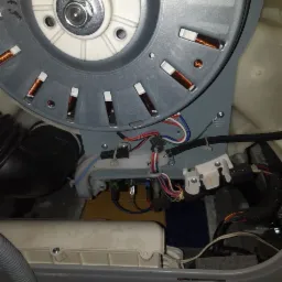 Wnętrze pralki z widocznym silnikiem, okablowaniem i elementami konstrukcyjnymi podczas naprawy. Widoczne złącza elektryczne i przewody o różnych kolorach.