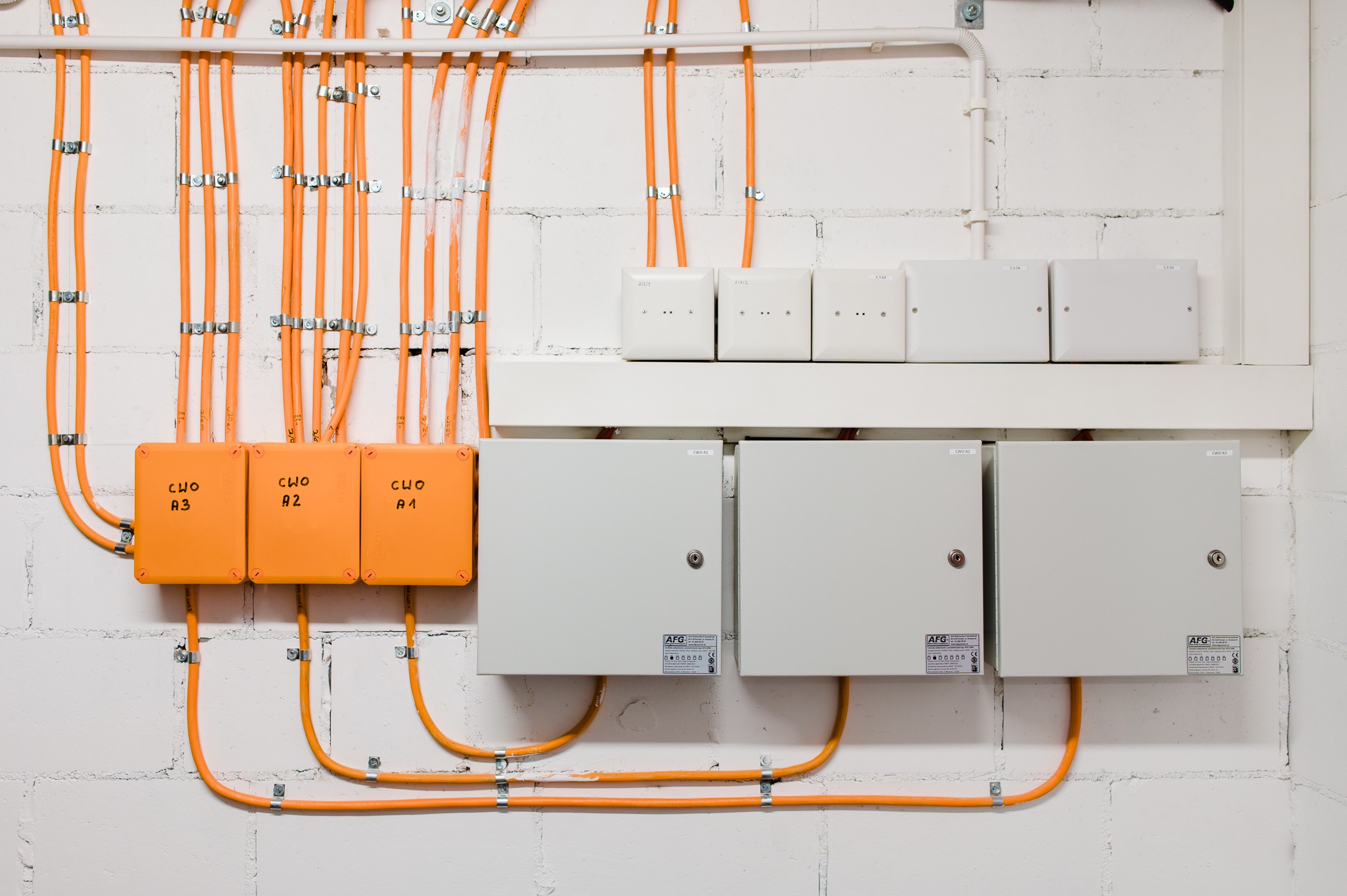 Pomarańczowe kable elektryczne przymocowane metalowymi uchwytami do ściany z białej cegły, prowadzące do trzech oznaczonych skrzynek CWO A1, A2, A3 oraz białych skrzynek kontrolnych i szafek...