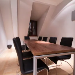 Sala konferencyjna w jasnym pomieszczeniu na poddaszu z długim drewnianym stołem na metalowych nogach i czarnymi krzesłami, widok na okno z rośliną.