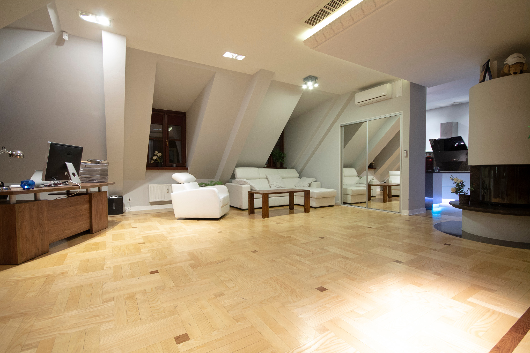 Przestronne wnętrze biurowe z drewnianą podłogą w jodełkę, białymi meblami wypoczynkowymi, biurkiem z komputerem i widoczną kuchnią w tle.