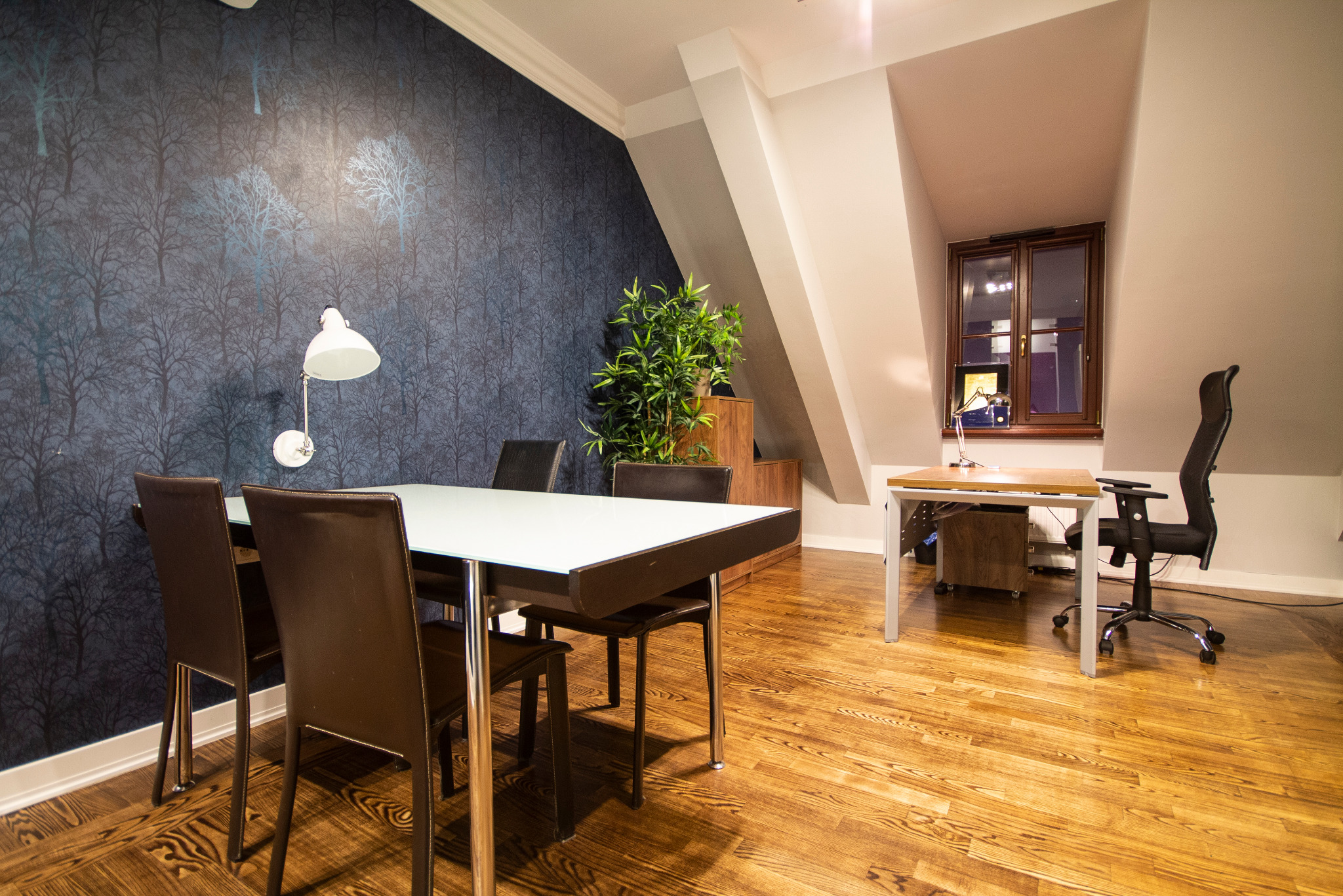 Przestronne biuro z drewnianą podłogą, dużym stołem konferencyjnym, krzesłami z brązowej skóry, rośliną doniczkową i ścianą w ciemnoniebieską tapetę z motywem drzew, oświetlone lampą biurkową