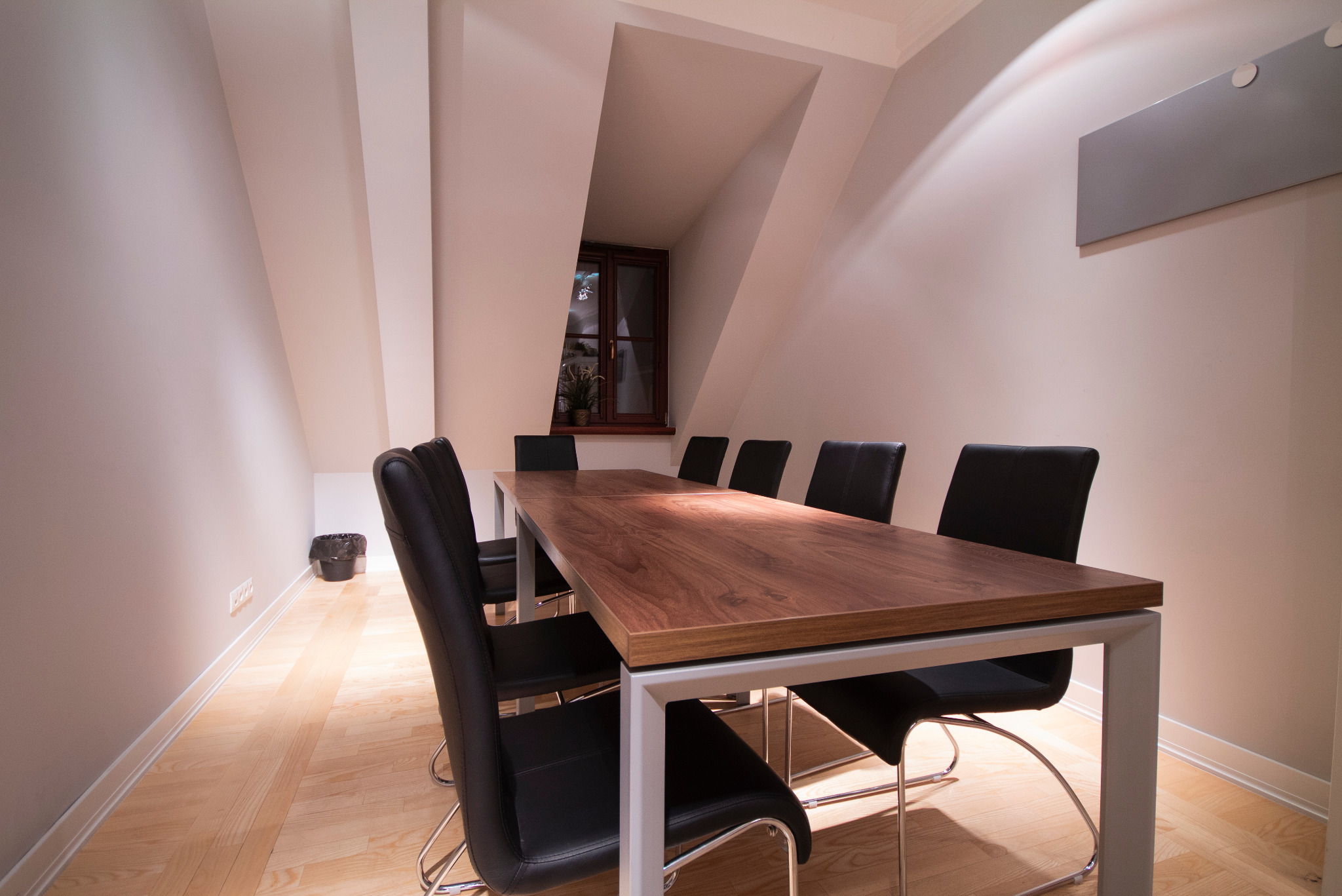 Sala konferencyjna w jasnym pomieszczeniu na poddaszu z długim drewnianym stołem na metalowych nogach i czarnymi krzesłami, widok na okno z rośliną.