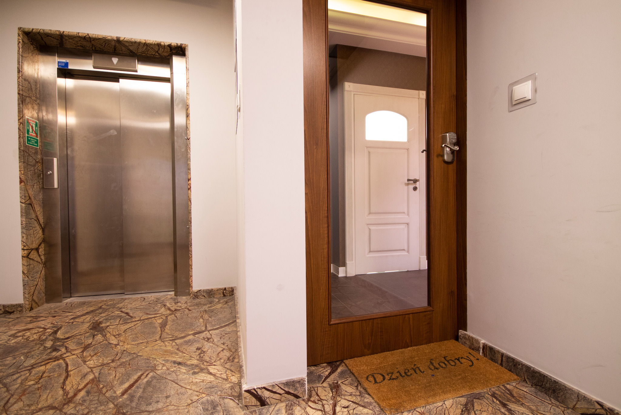 Wnętrze biurowca z windą o metalowych drzwiach w obudowie z marmuru i widocznym wejściem do biura z wycieraczką 'Dzień dobry!'