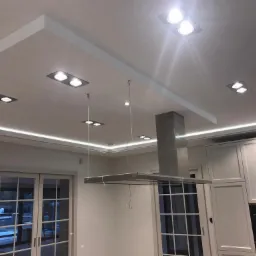 Nowoczesna kuchnia z oświetleniem LED podsufitowym, widoczne drzwi balkonowe i srebrny okap nad wyspą kuchenną.