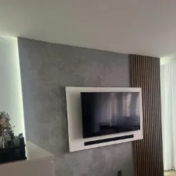Szara ściana wykończona dekoracyjnym tynkiem strukturalnym, z zamontowanym telewizorem w białej ramie i drewnianymi panelami pionowymi po prawej stronie, oświetlenie LED po lewej stronie.