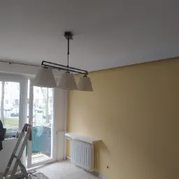 Pomalowane na żółto wnętrze pokoju z białym sufitem, wiszącą lampą z trzema abażurami i grzejnikiem, widoczne narzędzia malarskie i drabina, podłoga wyłożona panelami.