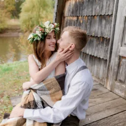 Uśmiechnięta para wpatrzona w siebie, kobieta z wiankiem na głowie, okryta kocem na tle drewnianej chaty i jeziora.