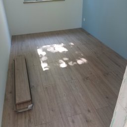 Firma Usługowa Złota Rączka Piotr Mikulewicz - Ułożone panele podłogowe w jasnym pokoju z oknem, z paczką paneli w rogu. Świeże, jasne wnętrze po remoncie. Widoczne promienie słoneczne na podłodze.