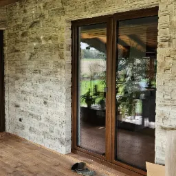 Zadaszony taras z kamienną okładziną ściany, widoczne duże przesuwne drzwi balkonowe z brązową ramą, drzwi wejściowe do wnętrza domu, meble ogrodowe i dekoracyjne rośliny doniczkowe.