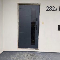 Stawski Okna - Grafitowe drzwi zewnętrzne z poziomymi panelami i pionowym przeszkleniem, zamontowane w surowym otworze, widoczny numer domu 282A na białej ścianie.