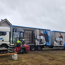 Stawski Okna - Pracownik w kamizelce odblaskowej obsługuje wózek widłowy Moffett, transportując palety z oknami z ciężarówki z reklamą firmy 'window 4u', na tle placu budowy z widocznym domem w trakcie budowy.