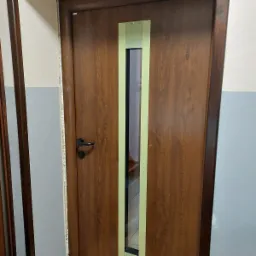 Brązowe drzwi wewnętrzne z wąskim przeszkleniem, zabezpieczone taśmą malarską, widoczne ślady montażu na ścianie.