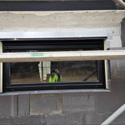 Stawski Okna - Montaż zewnętrznej rolety okiennej w trakcie prac budowlanych, widoczny fragment rusztowania i ocieplenie budynku grafitowym styropianem, odbicie osoby robiącej zdjęcie w szybie okna.