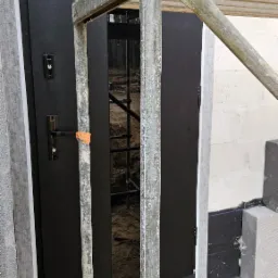Montaż czarnych drzwi zewnętrznych z widocznym fragmentem rusztowania i zabezpieczeniem przeciwwilgociowym przy fundamencie budynku.