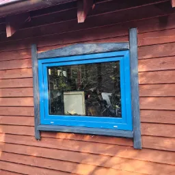 Błękitne okno PCV w drewnianej elewacji domu z widocznymi elementami ramy okiennej z czarnego drewna.