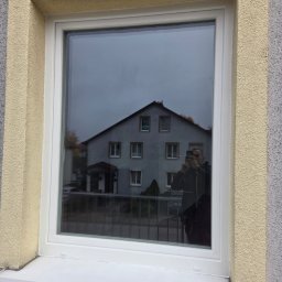 Stawski Okna - Białe okno PCV z widocznym odbiciem budynku i osoby robiącej zdjęcie. Żółta elewacja wokół okna. Nowoczesny design.