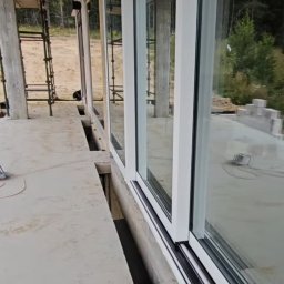 Stawski Okna - Białe, przesuwne okna PCV zamontowane w surowym budynku, widoczne z zewnątrz. Na pierwszym planie betonowa podłoga z otworami. W tle las i rusztowanie.