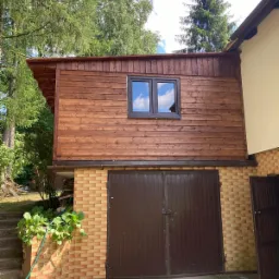 Garaż z brązową bramą dwuskrzydłową, obok wejście do budynku, nad garażem drewniana nadbudowa z oknem, całość otoczona zielenią.