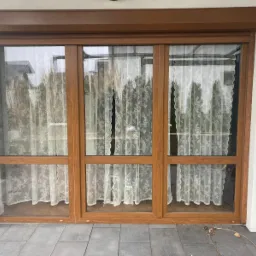 Trzyczęściowe przesuwne okno z brązowymi ramami i zewnętrzną roletą, widoczne firanki w oknach, szare płytki na zewnątrz, różowe klapki leżące obok okna.