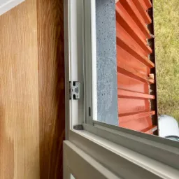 Detal uchylnego okna z widocznym mechanizmem zawiasu, drewnianym panelem wewnętrznym i kroplami deszczu na szybie zewnętrznej.