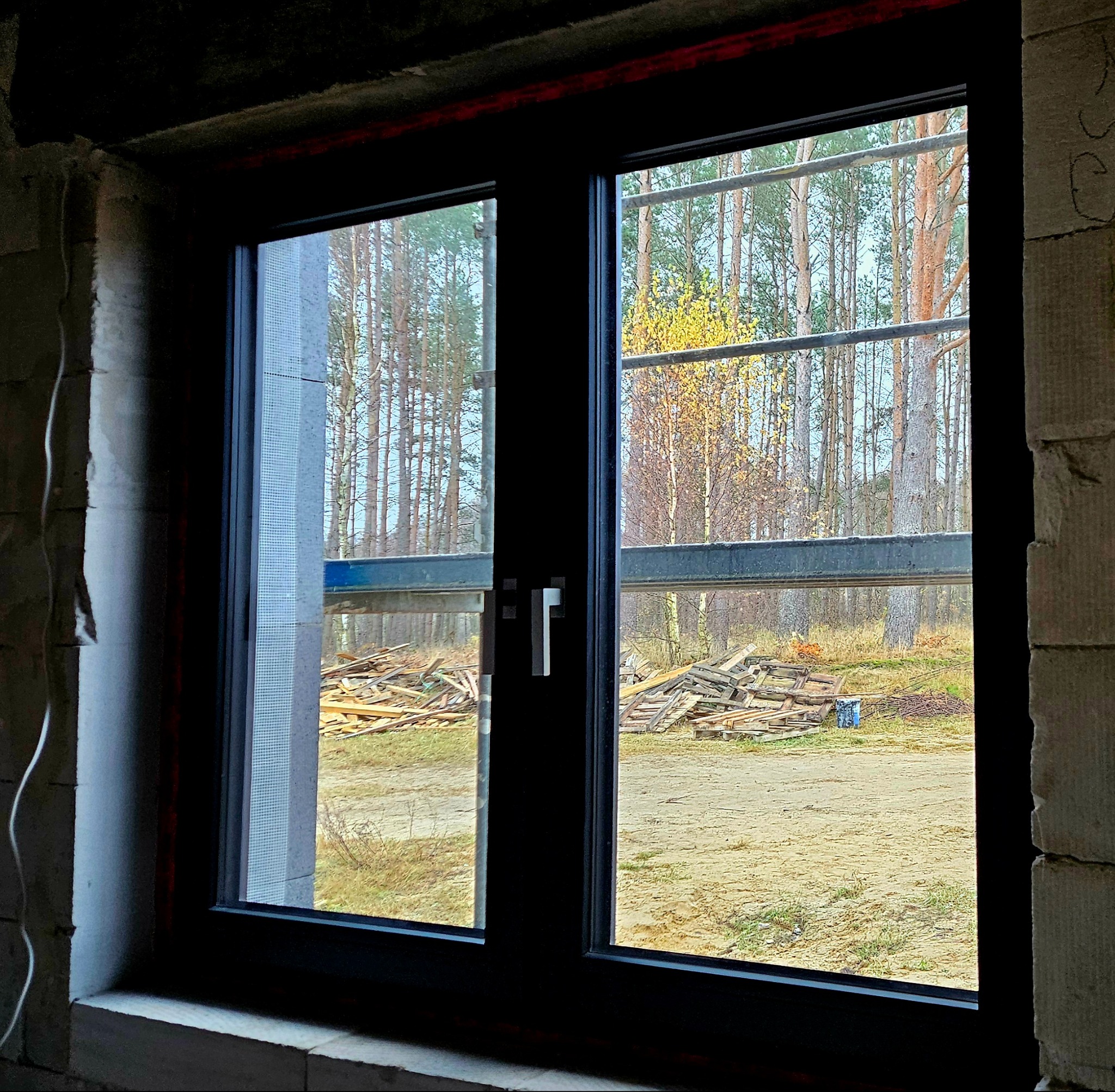 Czarne dwuskrzydłowe okno z widokiem na las w jesiennej scenerii, widoczne rusztowanie i stos desek na zewnątrz, surowy stan budynku wewnątrz.
