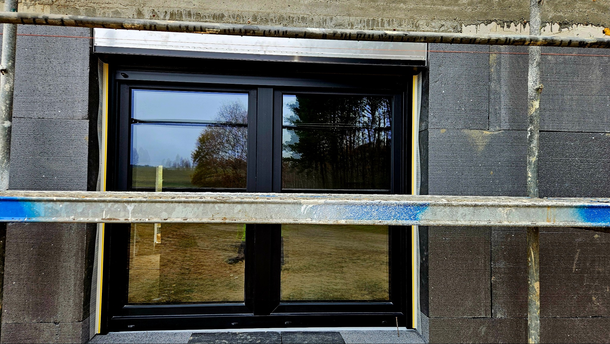 Czarne okno PCV z roletą zewnętrzną w trakcie montażu, widoczne rusztowanie i ocieplenie elewacji styropianem grafitowym.