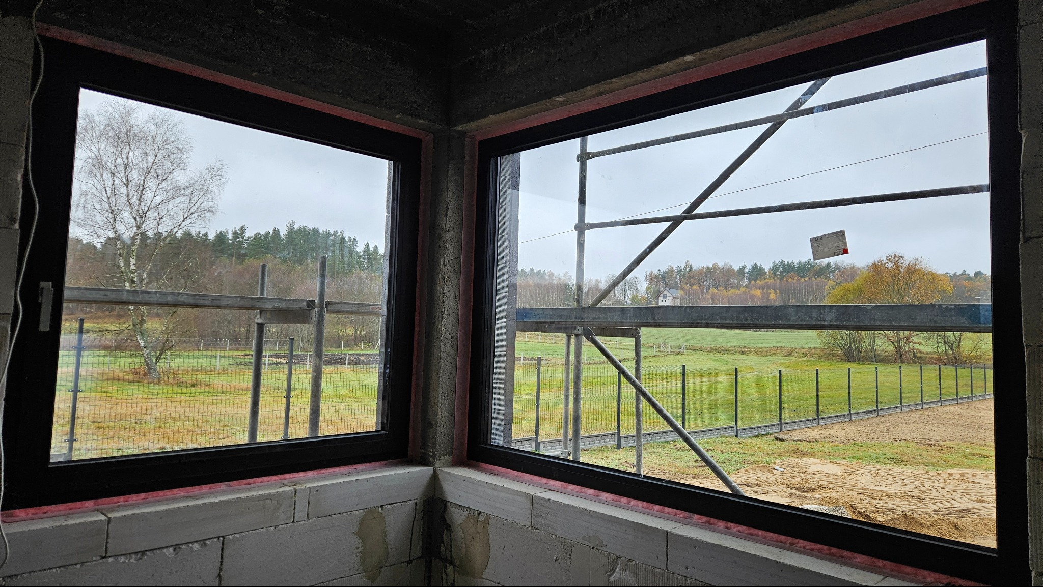 Dwa czarne okna w surowym stanie, widok na zewnątrz na łąkę, las i rusztowania, z bliska widoczny mur z pustaków.