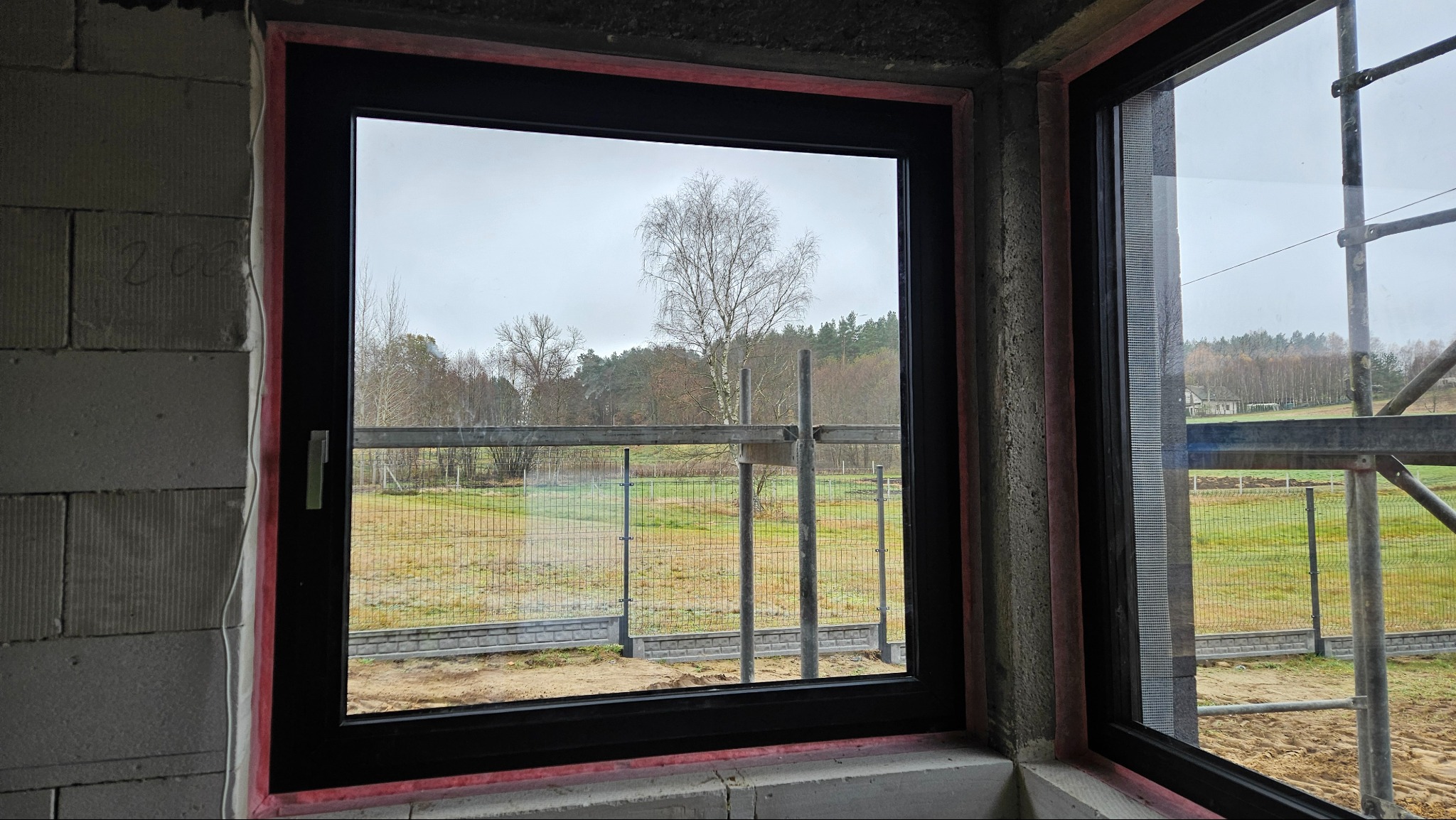 Czarne okno w surowym stanie, widok na zewnątrz z rusztowaniem i drzewami w tle, czerwona pianka montażowa wokół ramy.