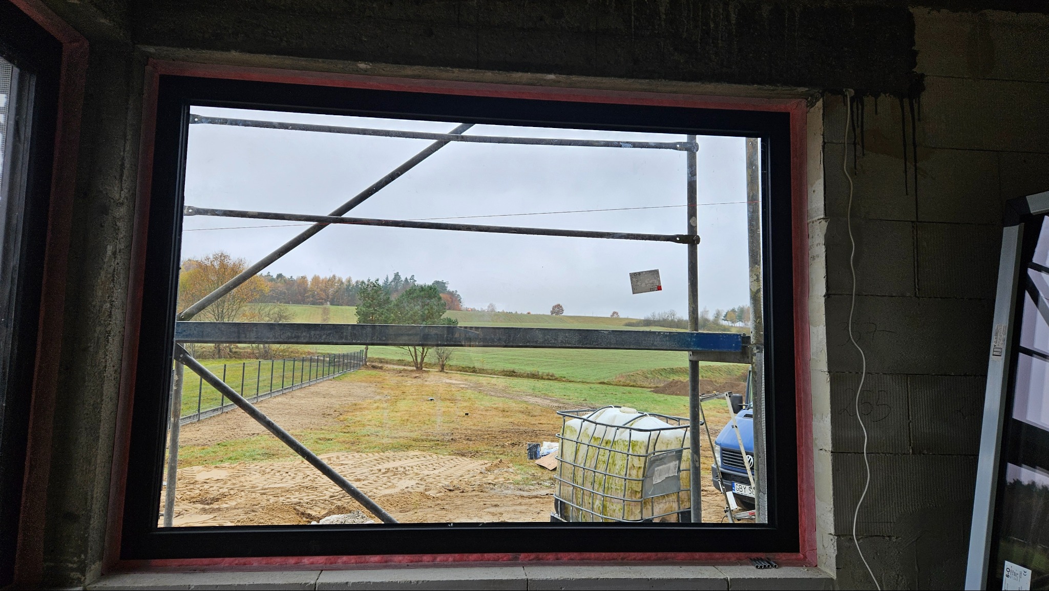 Surowy montaż okna z czarną ramą w ścianie z betonu komórkowego, widok na krajobraz z rusztowaniem i zbiornikiem IBC na zewnątrz.