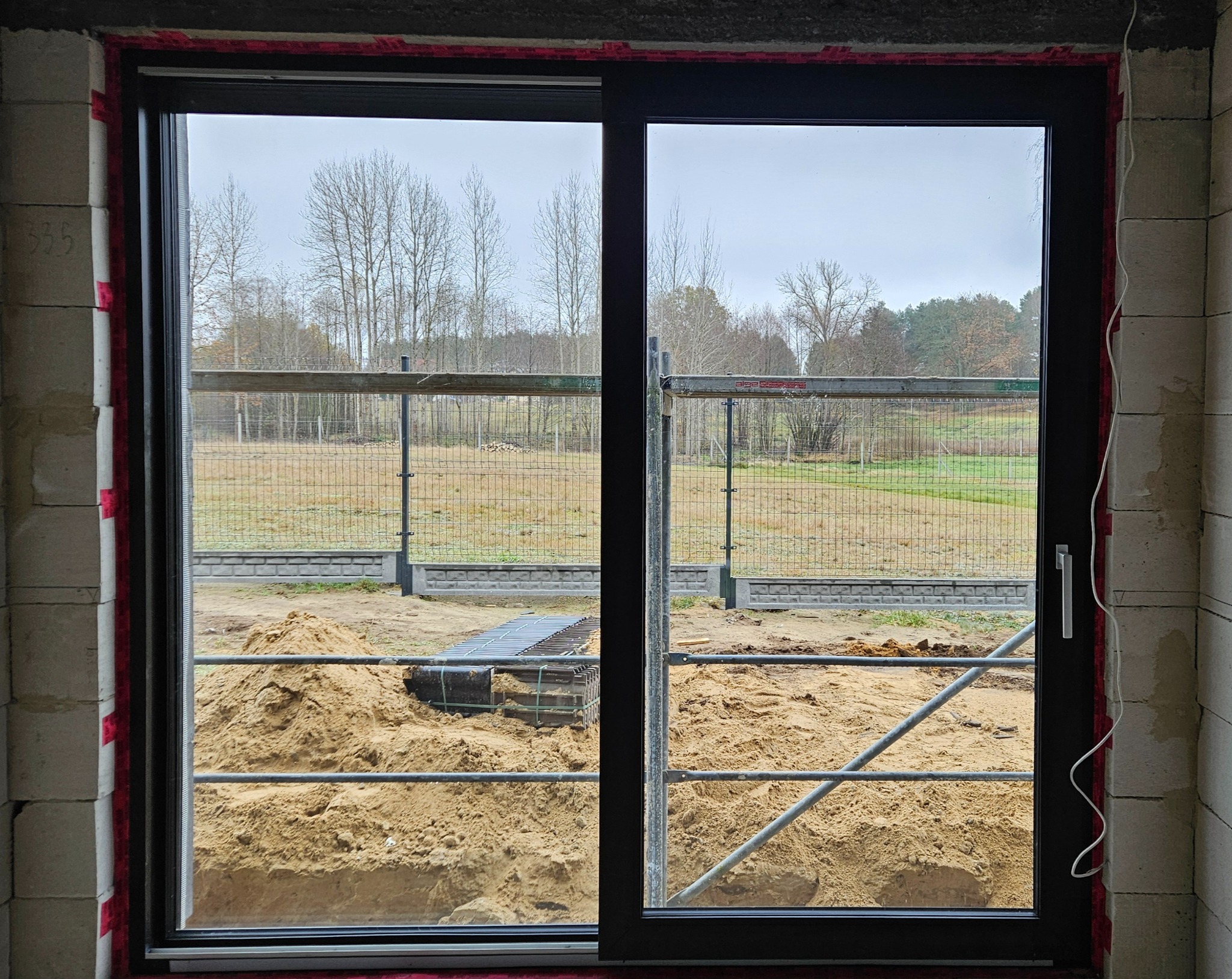 Czarne przesuwne okno w surowym stanie, widok na plac budowy z piaskiem i ogrodzeniem w tle.