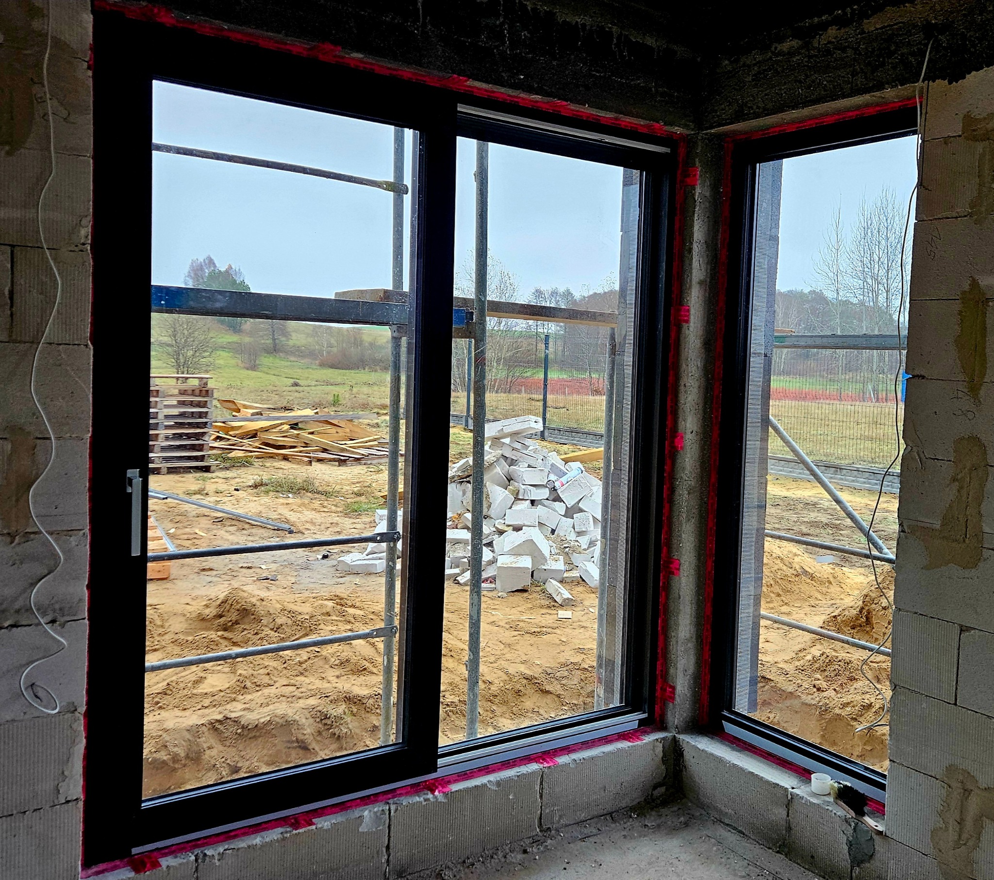 Surowe wnętrze w trakcie budowy z zamontowanymi czarnymi oknami, widok na plac budowy z rusztowaniami i stertą gruzu za oknem.