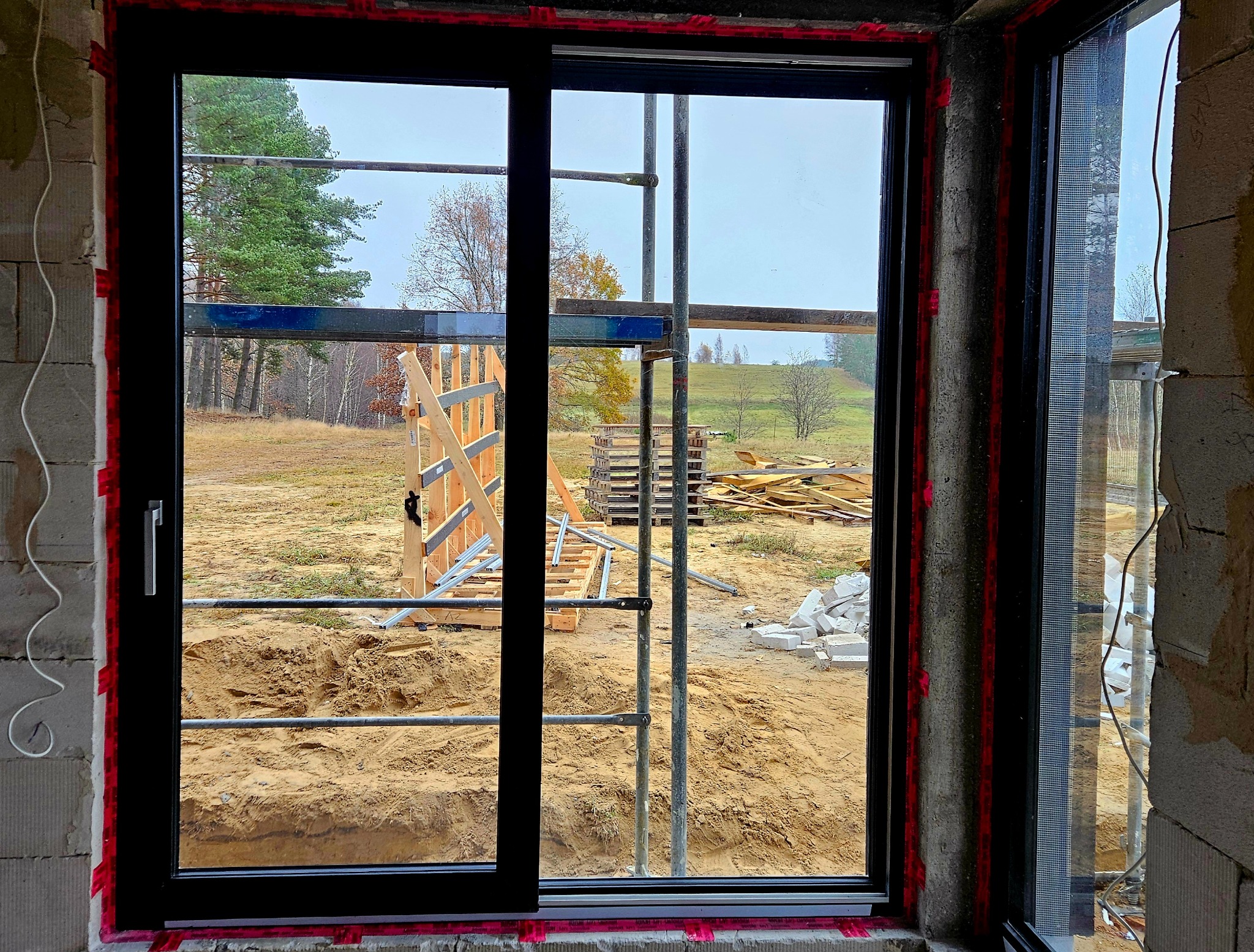 Czarne okno przesuwne z widokiem na plac budowy, widoczne rusztowania, drewniane konstrukcje i stosy materiałów budowlanych na zewnątrz.