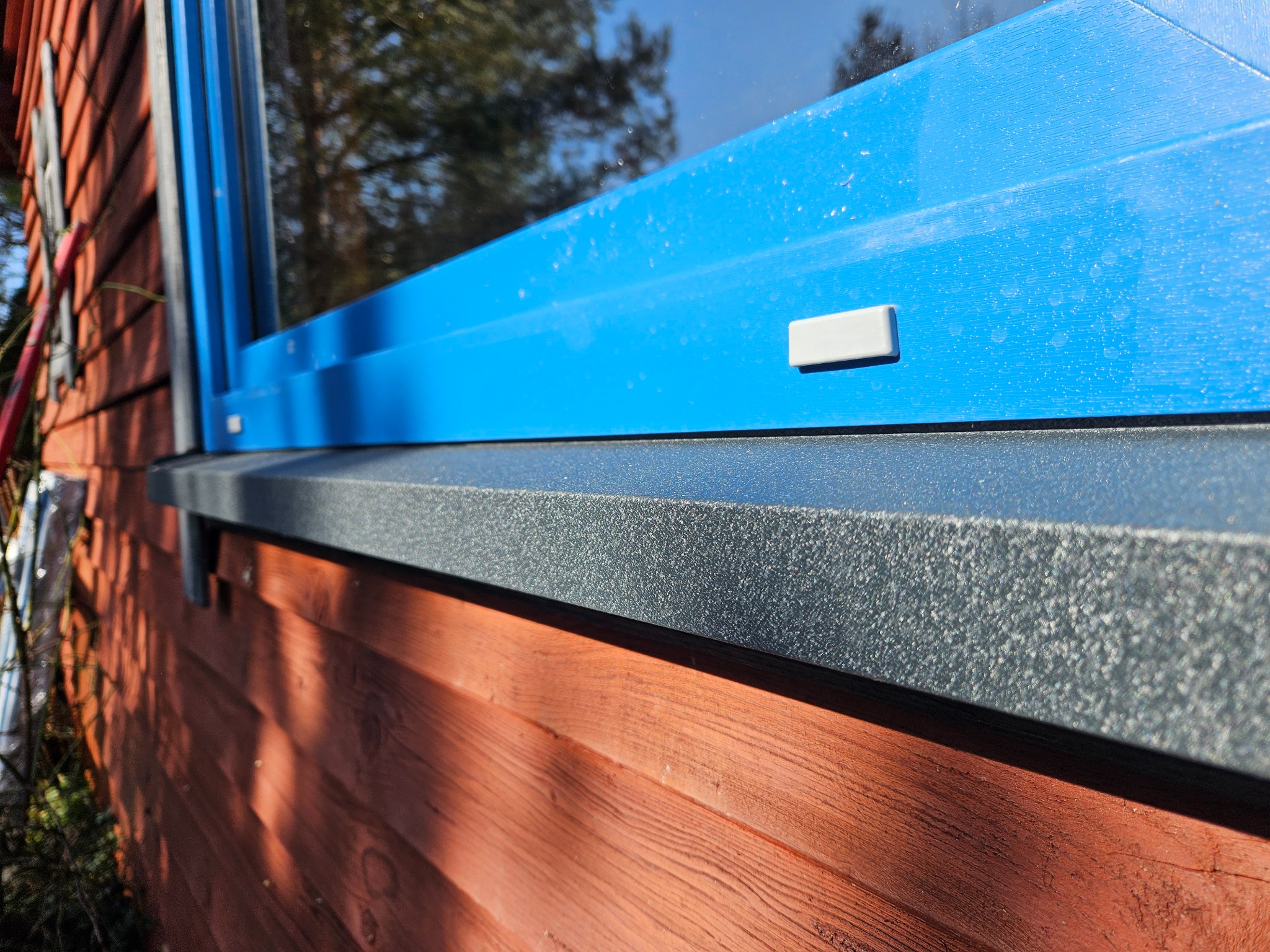 Niebieska rama okienna z szarym parapetem zewnętrznym zamontowana na drewnianej elewacji domu, widoczne odbicie drzew w szybie.