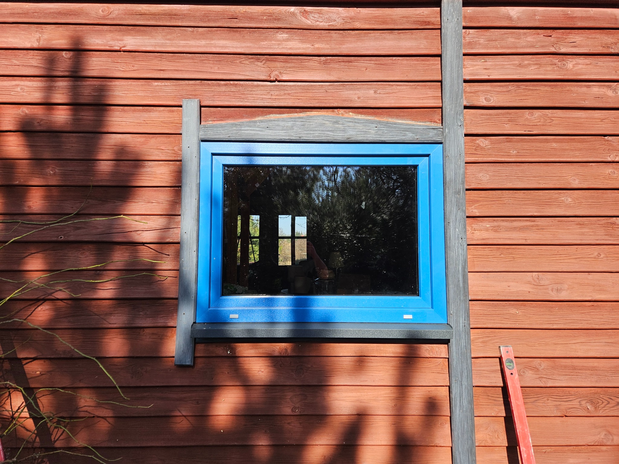 Błękitna rama okienna na tle drewnianej ściany z widocznym poziomem i refleksami drzew w szybie.